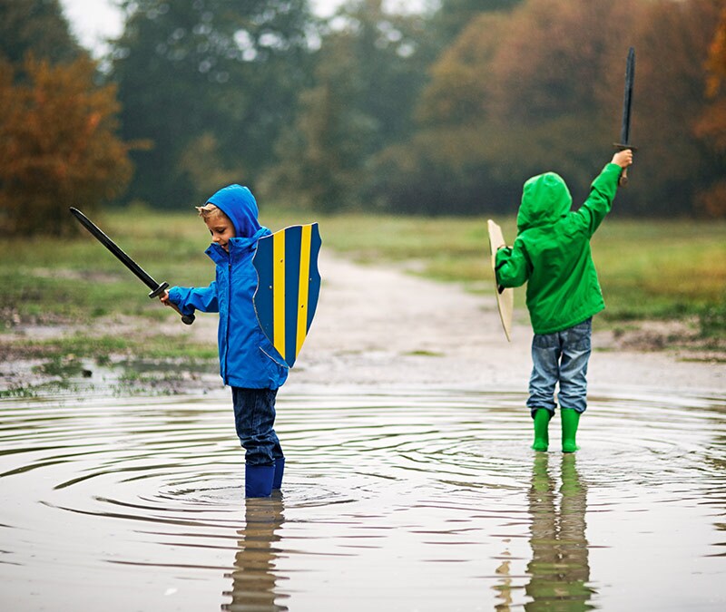 Two children standing in a puddle wearing rain gear and holding toy swords and shields.