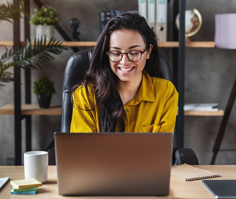 A woman wearing glasses smiles as she uses a laptop.